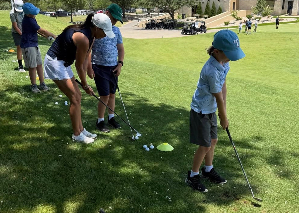 A family golfing experience on a sunny day at a golf course, with a woman and a young boy in golf attire preparing to hit golf balls on the driving range.