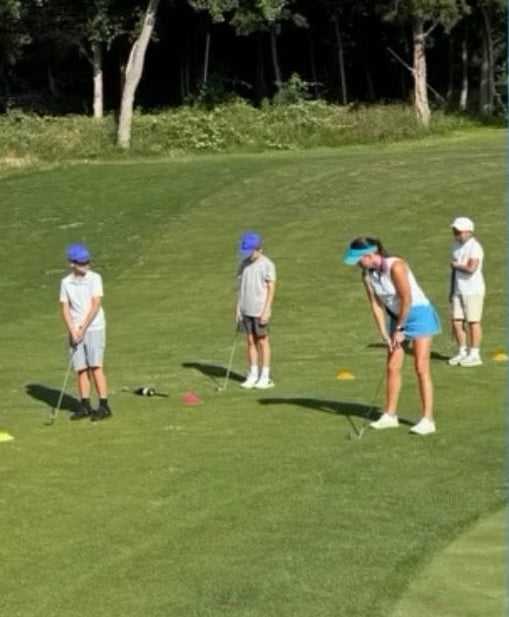 Outdoor golf course scene with several people playing on a grassy fairway under a blue sky, with trees in the background. Large overlaid text reads “SUMMER GOLF CAMPS,” “#ni kegolfcamp,” and “Cowboys Golf Club.